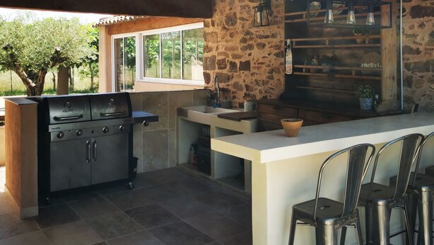 An outdoor kitchen area with a stone wall, a grill, bar stools, and a view of the surrounding landscape.