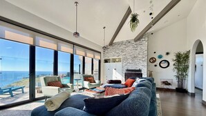 Una sala de estar moderna con un sofá grande, una mesita de centro y vistas al océano. Hay dos sillones, una chimenea y un ventanal amplio.