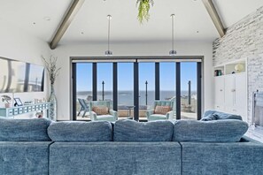 Un salon moderne doté d’un grand canapé, de deux fauteuils, d’une table basse et offrant une vue sur l’océan grâce à de grandes fenêtres du sol au plafond.