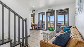 Un interior moderno con una escalera, una zona de estar con sofá y cojines, una mesa de comedor y ventanas amplias con vistas al mar.