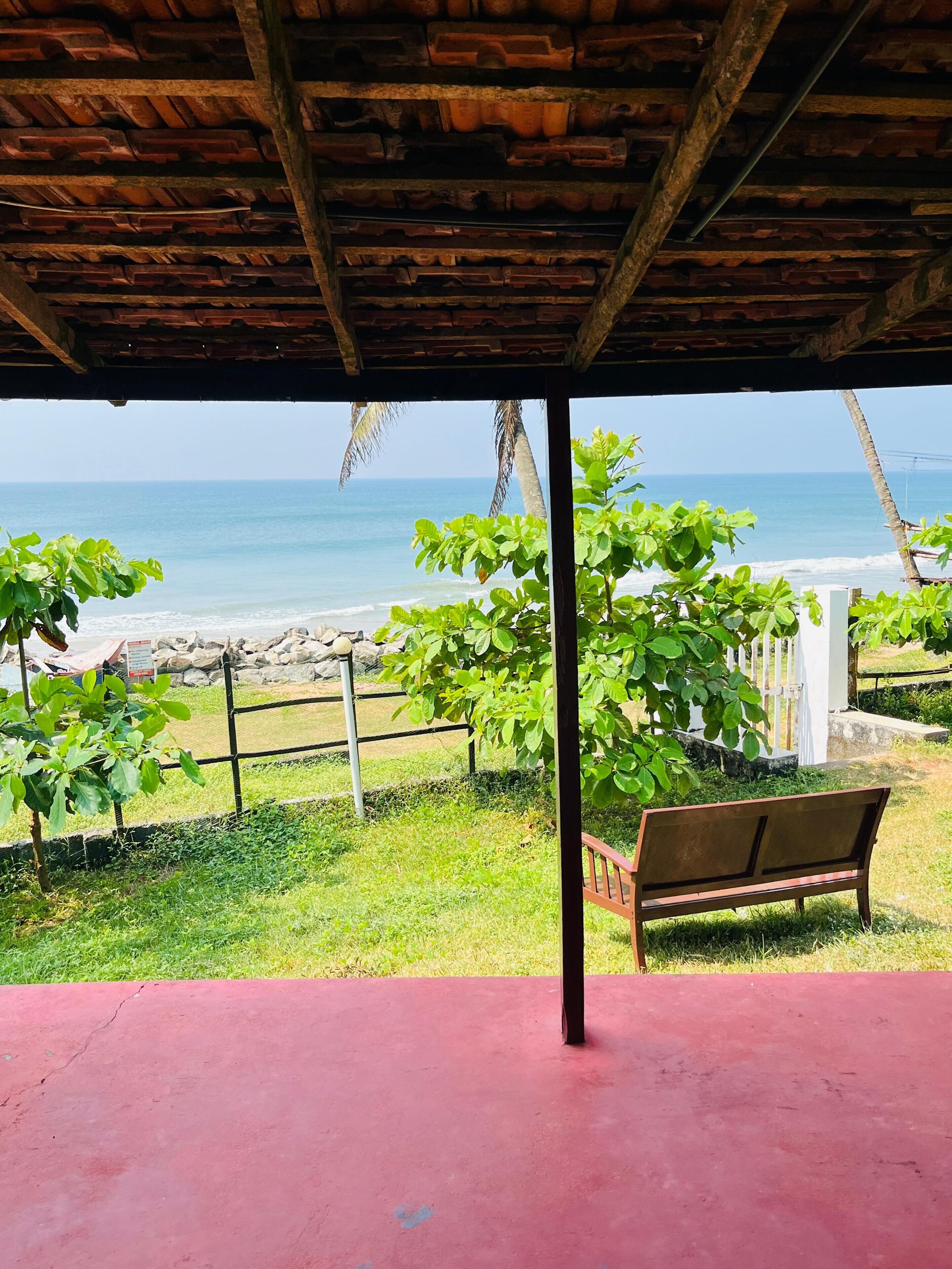 Uma área externa coberta com um banco, vista para a praia e uma palmeira.