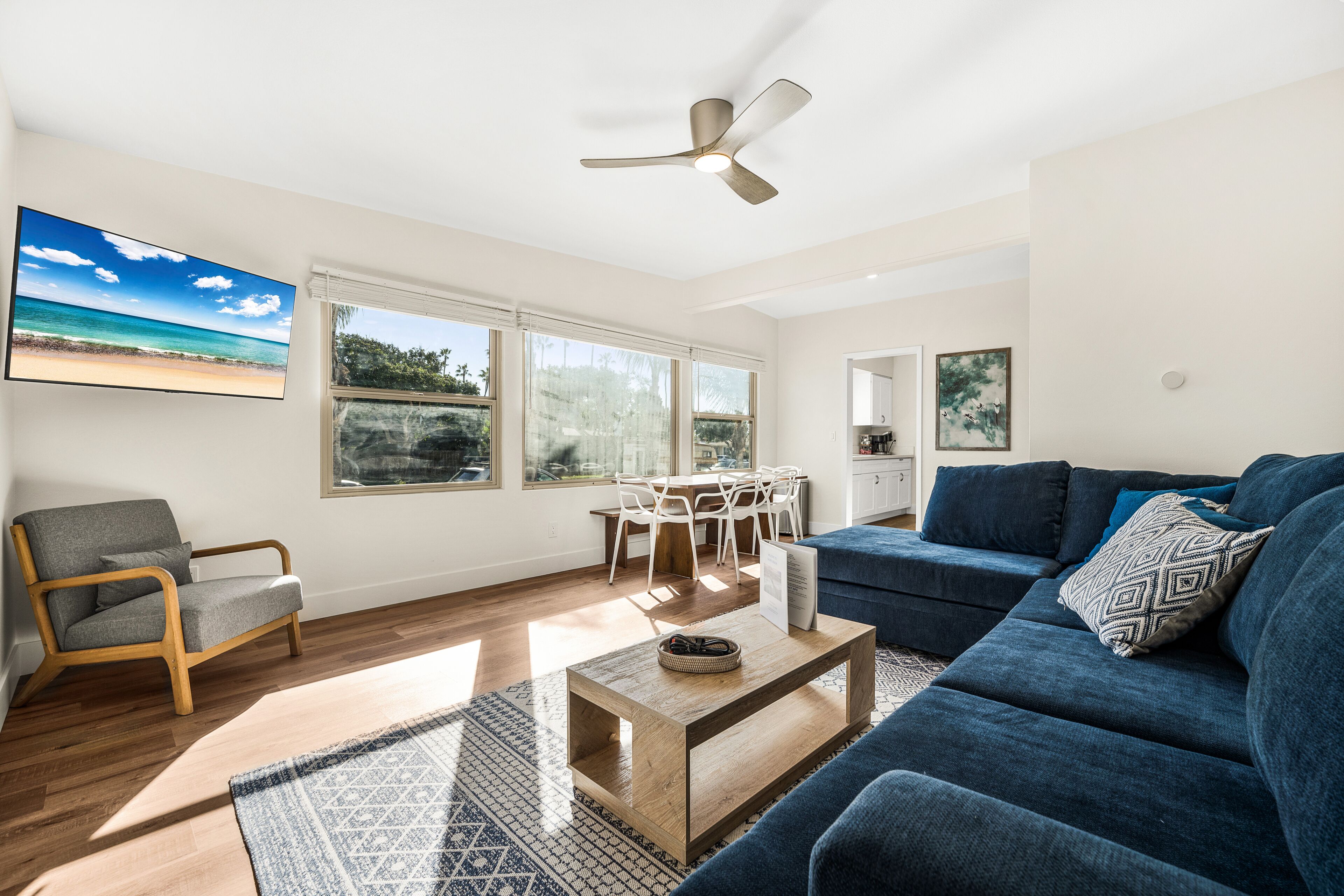 A spacious living room with a blue sofa, a wooden coffee table, and a grey armchair. There is a dining area with white chairs and a table, and a ceiling fan.