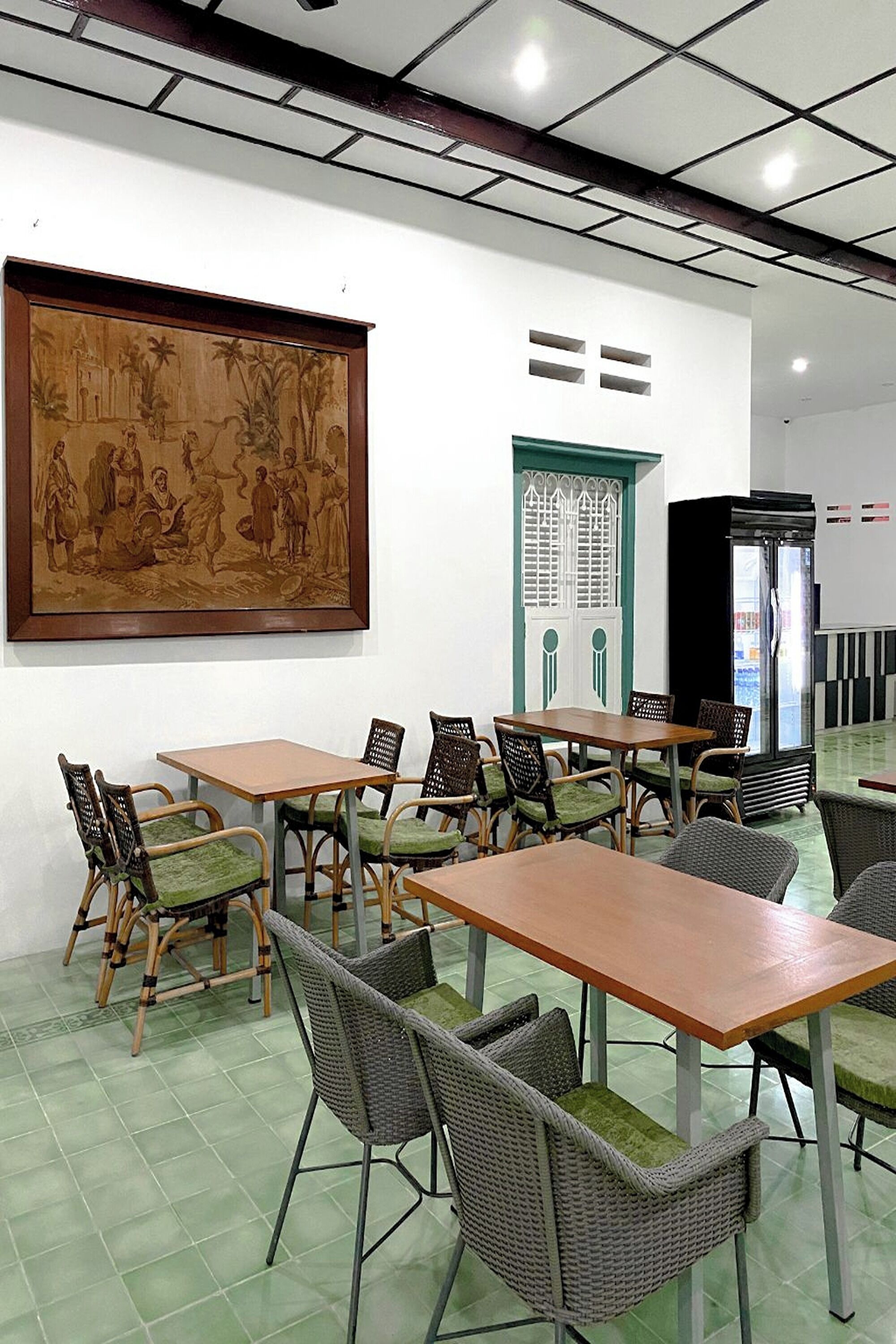 A dining area with tables and chairs, a large framed artwork on the wall, and a refrigerator in the background.