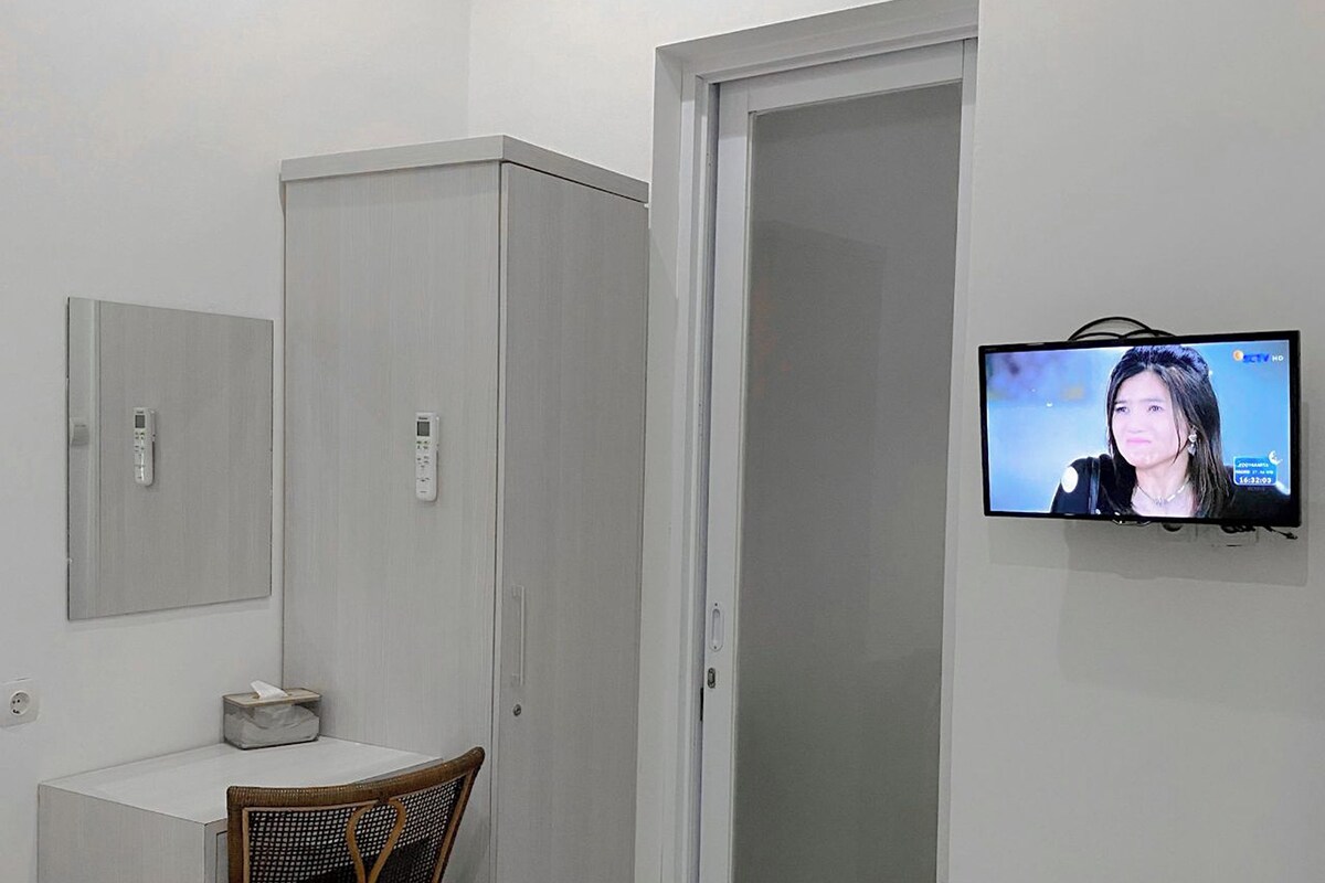 A modern room with a white wardrobe, a small table with a tissue box, a wicker chair, and a TV mounted on the wall.