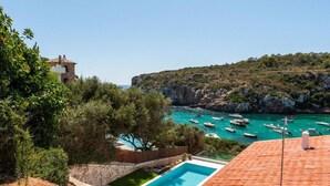 Uma villa costeira com piscina, falésias rochosas e barcos ao fundo.