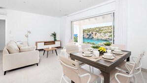 Uma sala de estar moderna com área de jantar, móveis brancos, um armário de madeira e uma grande janela com vista para a costa.