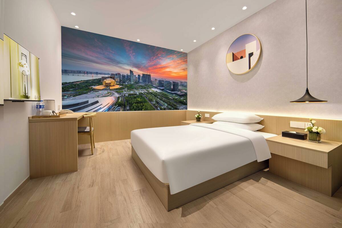 Habitació d'hotel moderna amb un mural gran de paisatge urbà, un llit ben fet, una taula de treball amb una cadira i una tauleta de nit amb una làmpada.