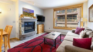 A living room with a stone fireplace, a flat-screen TV, a wooden dining table, and a red patterned rug. - 8045 Black Bear 1 Bedroom Condo by RedAwning (Keystone)