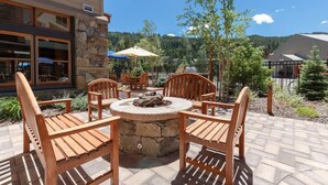An outdoor patio with a stone fire pit surrounded by wooden chairs, a stone wall, and a view of a forested mountain range. - 8045 Black Bear 1 Bedroom Condo by RedAwning (Keystone)