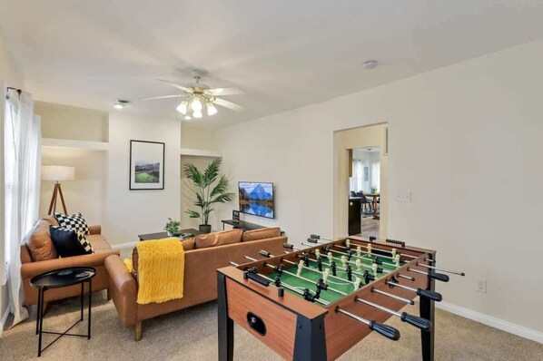 A living room with a foosball table, a brown sofa, a yellow throw blanket, a ceiling fan, and a framed picture on the wall.