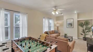 A living room with a foosball table, a ceiling fan, a sofa, a coffee table, and a potted plant.