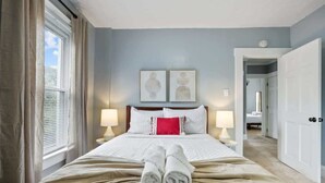 A hotel room with a bed, two wall-mounted lamps, a window with curtains, and a door leading to another room. - The Indiana Stunner S (Columbus)