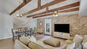 Una sala de estar con pared de ladrillo, vigas de madera, comedor con mesa y sillas, y televisor sobre un mueble.
