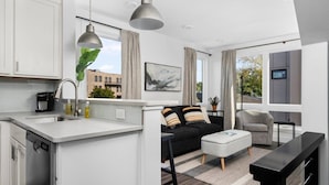 A modern kitchen with white cabinets, a built-in dishwasher, and a large island with a sink. There is a black sofa with striped cushions and a grey armchair. A large window provides a view of the city.