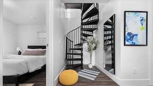 A modern bedroom with a bed, a yellow pouf, a striped rug, a black spiral staircase, and a framed abstract painting.