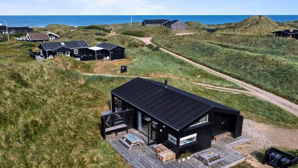 A modern black building with a deck, surrounded by grassy dunes and a clear view of the sea.