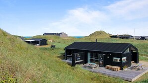 A modern black building with a wooden deck, surrounded by grassy dunes and other structures.