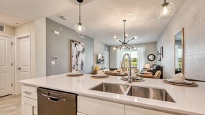 Una cocina moderna con una isla central, electrodomésticos de acero inoxidable, iluminación colgante y vistas al salón.