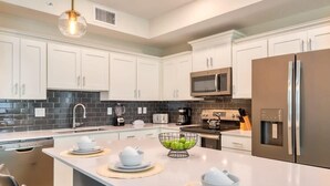 A modern kitchen with white cabinets, a stainless steel refrigerator, a microwave, and a large island with a bowl of fruit.