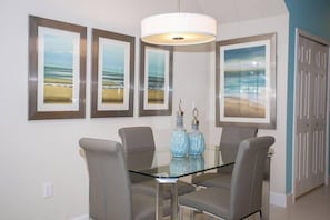 Un comedor con una mesa de vidrio y cuatro sillas, con pinturas de playa en la pared y una puerta a la derecha.
