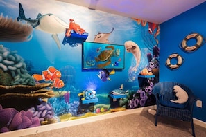 Une pièce avec une grande fresque représentant un paysage sous-marin, un téléviseur fixé au mur et un fauteuil bleu.