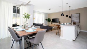 Uma sala de estar moderna com sofá, mesa de jantar posta para quatro e área de cozinha com televisão e luminárias pendentes.