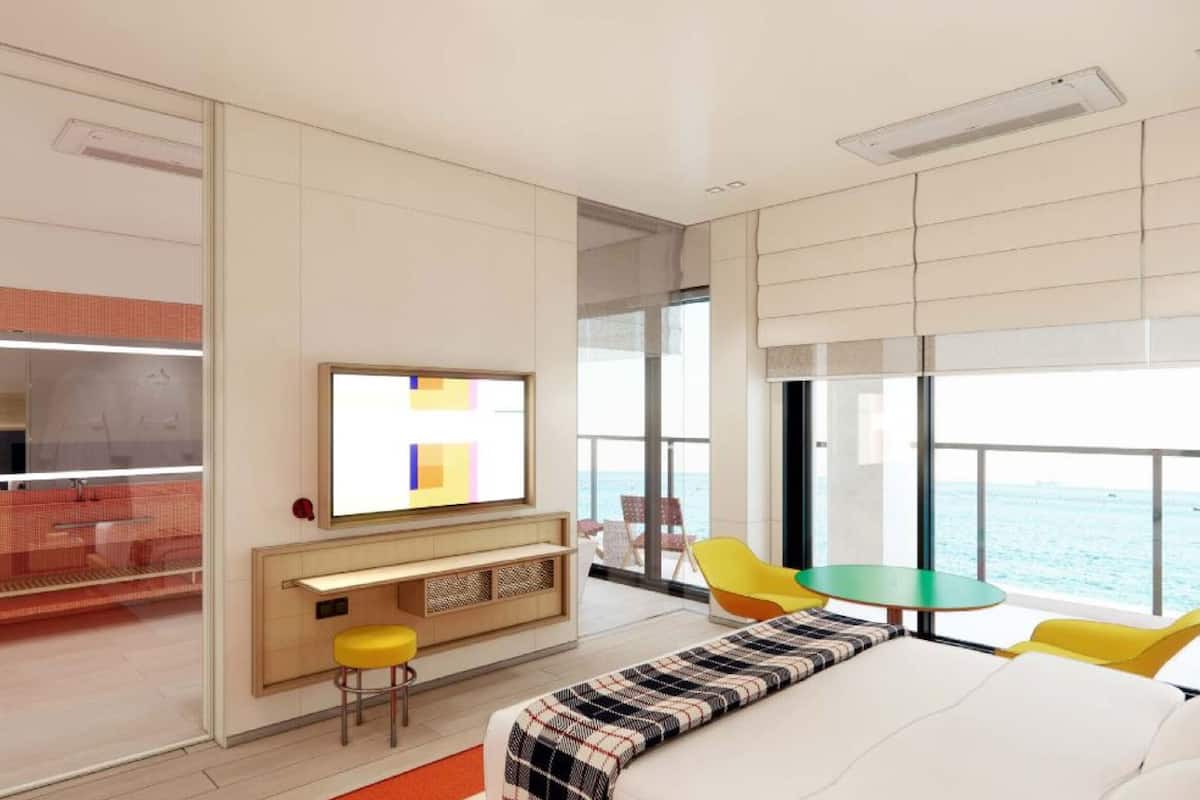 Una habitación de hotel moderna con una cama grande, televisión, zona de estar con mesa y sillas, y vista al mar.