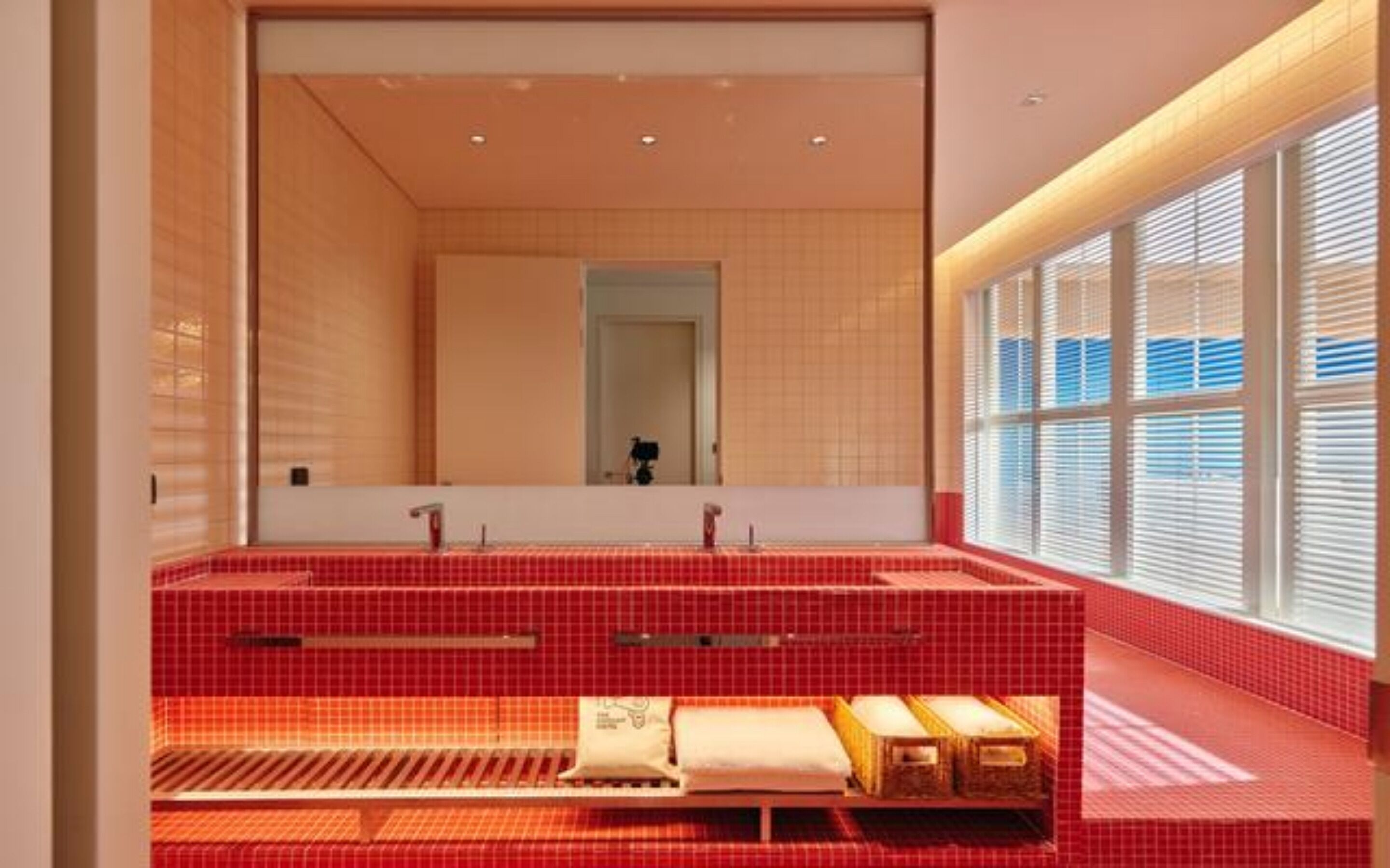 Ein modernes Badezimmer mit roten Fliesen, einem großen Spiegel und einem Fenster mit Jalousien.