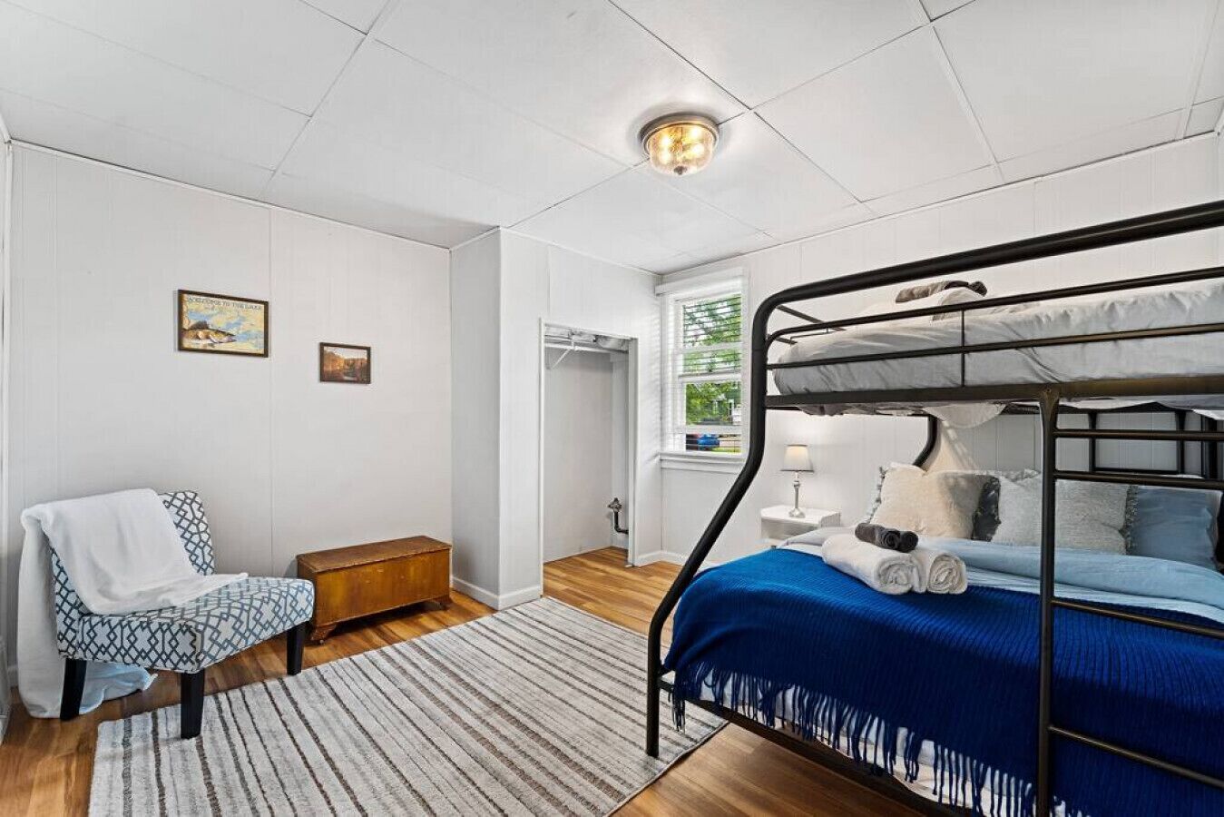 A bedroom with a bunk bed, a chair, a small wooden chest, and a window with a view of greenery.
