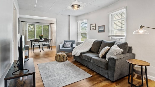 A modern living room with a grey sofa, a coffee table, and a TV on a stand. There is a dining area with a table and chairs, and a window with blinds. - Pattison`s Peaceful Retreat (Superior)