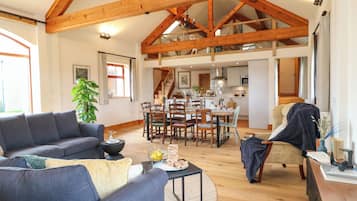 A spacious living area with a high ceiling and exposed wooden beams, a dining area with a wooden table and chairs, and a kitchen in the background.