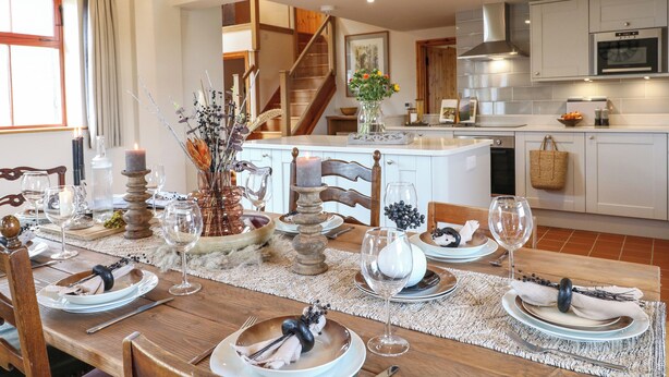 A dining area with a wooden table set for a meal, a kitchen in the background, and a staircase leading to an upper level. - Rowtor (Matlock)