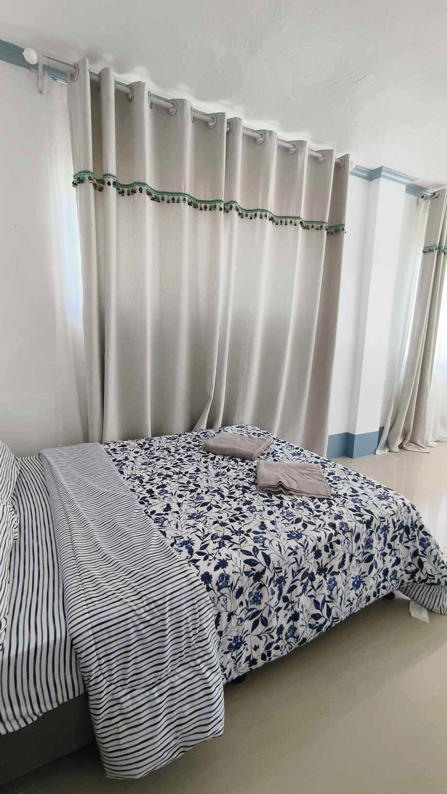 Een bed met gestreepte en bloemenprints, een gordijn met groene biezen, en een effen muur.