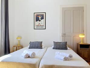 Een hotelkamer met twee bedden, elk met een opgerolde handdoek, een ingelijste poster aan de muur en bedlampjes.