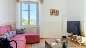 Ein Wohnzimmer mit einem roten Sofa, einem Fernseher, einem Holztisch und einem Fenster mit Blick auf Bäume.