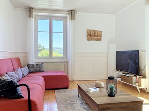 Een woonkamer met een rode bank, een televisie, een houten bijzettafeltje en een raam met uitzicht op bomen.