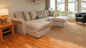 A living room with a beige sectional sofa, a small ottoman, a wooden coffee table, and a lamp. There are framed pictures on the wall and a view of the outside through a glass door. - Cullernose Cottage (Alnwick)