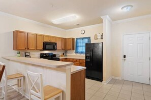 A kitchen with wooden cabinets, a black refrigerator, a microwave, and a sink. There is a white door and a window with blue curtains.