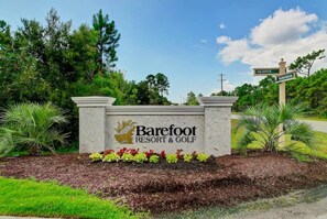 A sign for Barefoot Resort & Golf with a logo, surrounded by greenery and a clear sky.