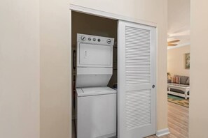 Une petite buanderie équipée d’un lave-linge et d’un sèche-linge intégrés, d’une porte blanche à panneaux ajourés et d’un petit coin salon visible à travers une porte ouverte.
