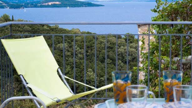 Un balcón con una silla verde, una mesa con vasos y una vista panorámica de un lago y montañas.