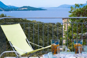 A balcony with a green chair, a table with glasses, and a scenic view of a lake and mountains. - Holiday Apartment "Scorpios View" with Sea View, Private Terrace & Wi-Fi (Meganisi)