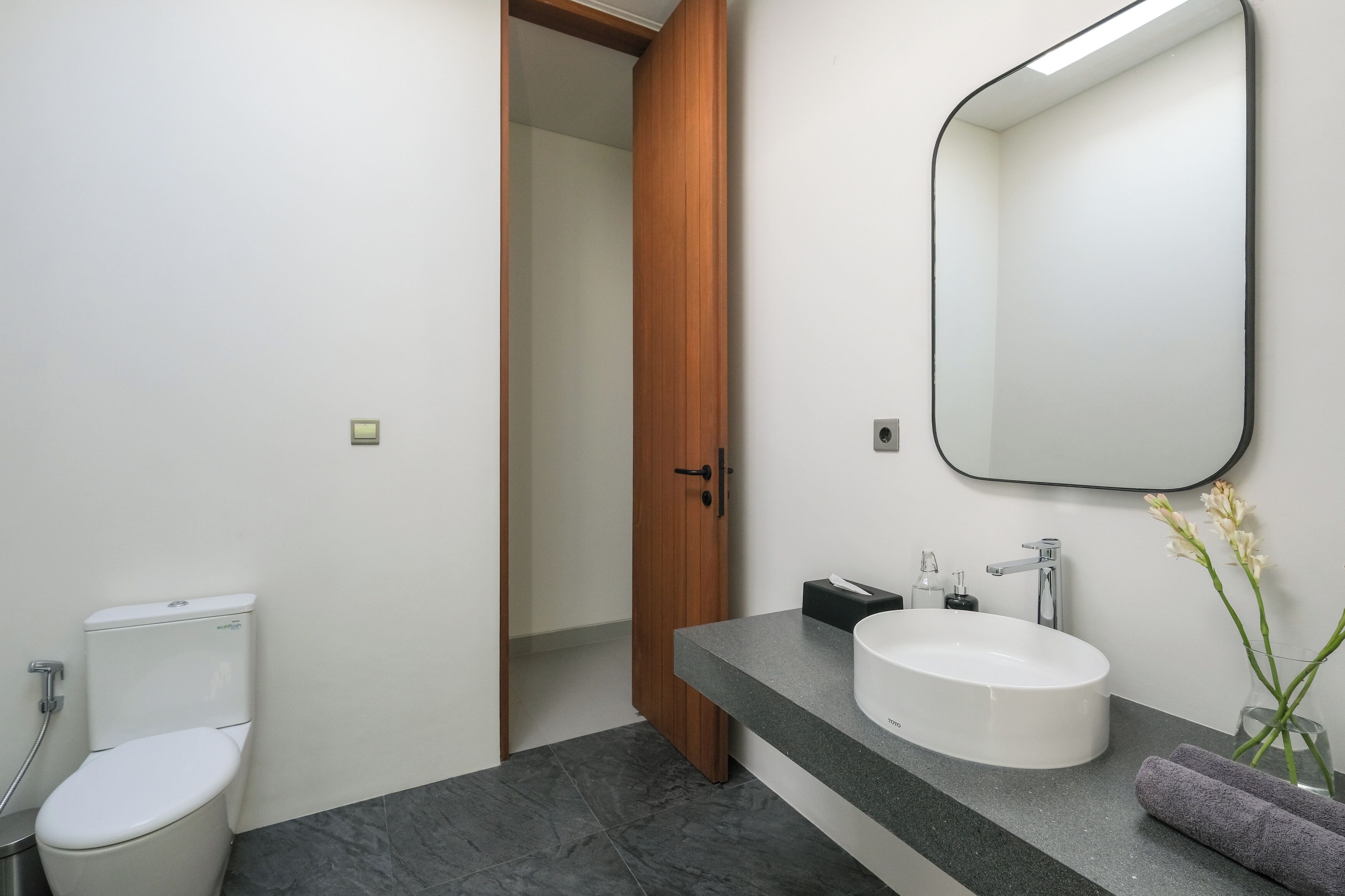 A modern bathroom with a white toilet, a wooden door, a large mirror, and a granite countertop with a sink.