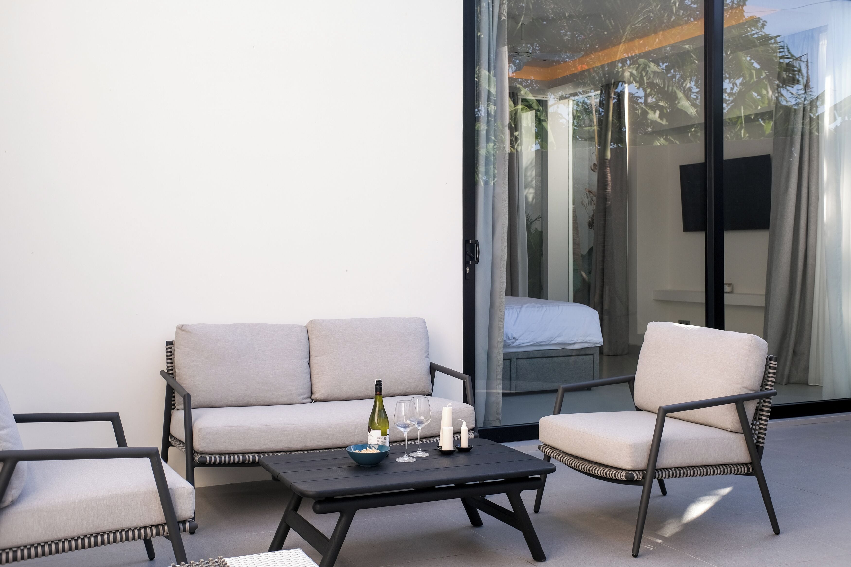 A modern outdoor seating area with a sofa and two chairs, a table with a bottle and glasses, and a glass door leading to a balcony.