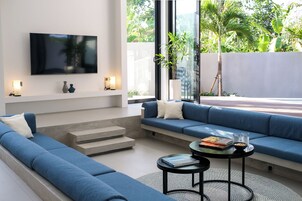 Una sala de estar moderna con un sofá azul, un televisor de pantalla plana montado en la pared y un ventanal con vistas a la vegetación.