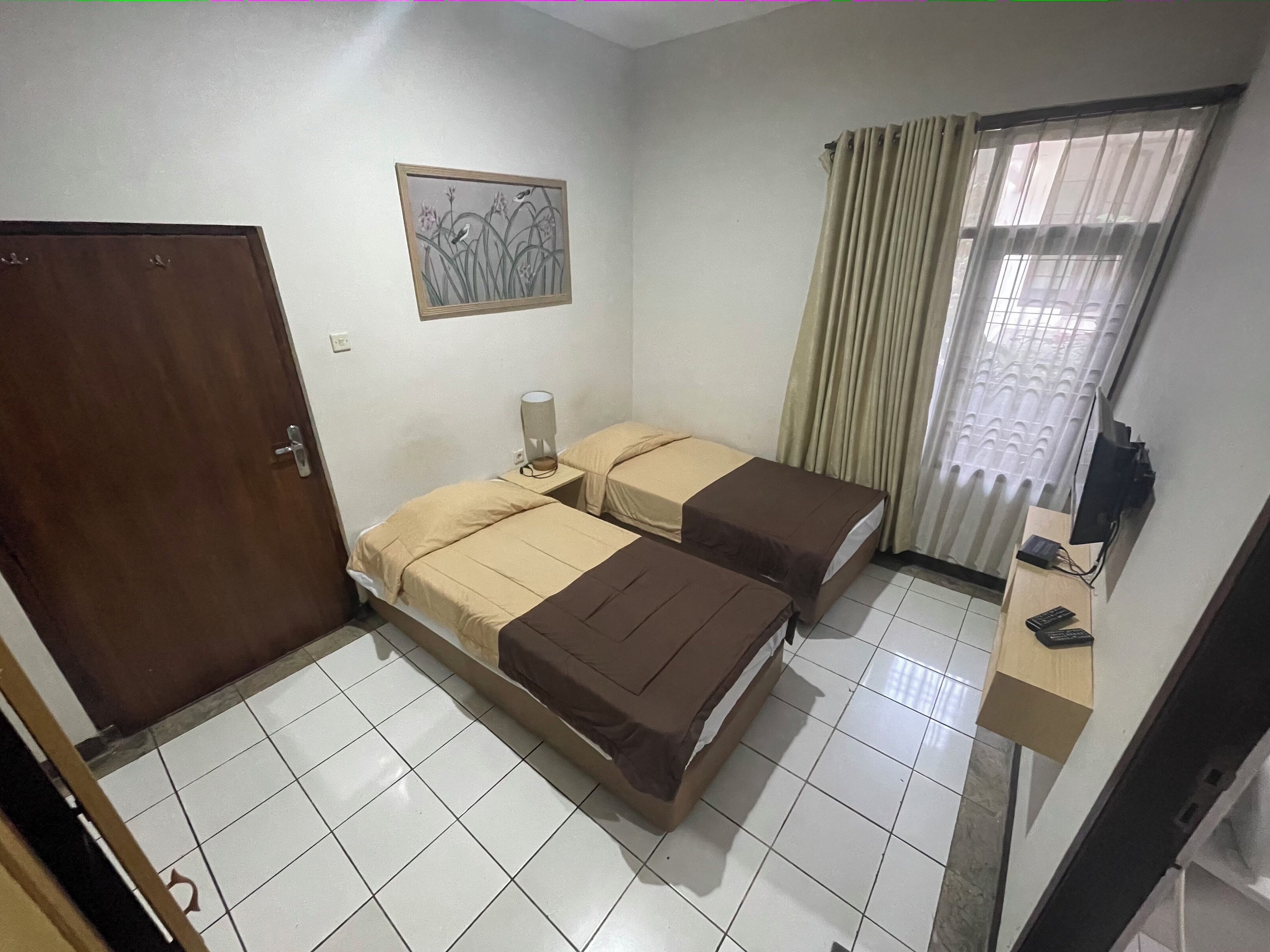 A small, clean room with two beds, a door, a painting, a window with curtains, and a desk with a lamp.