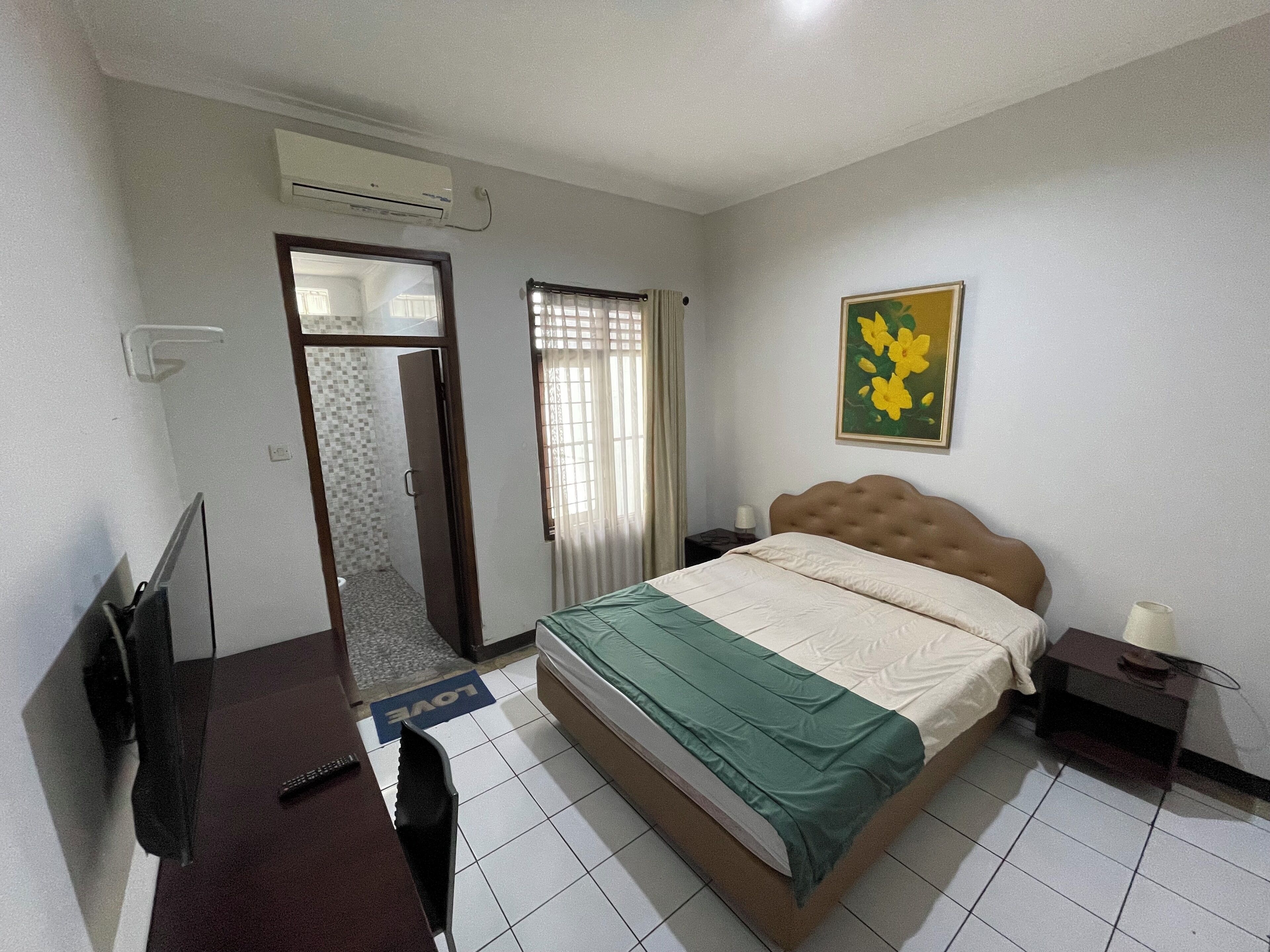 A bedroom with a bed, a desk, a chair, an air conditioning unit, a painting, and a door leading to a bathroom.