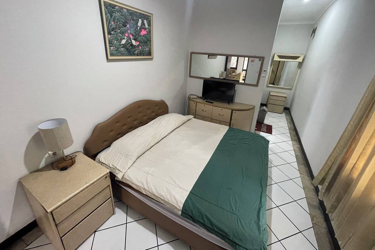 A bedroom with a bed, bedside table, lamp, dresser, TV, and a painting on the wall.