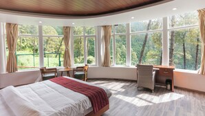 Quarto com uma cama grande, uma escrivaninha e uma cadeira, com vista para árvores através de janelas amplas.