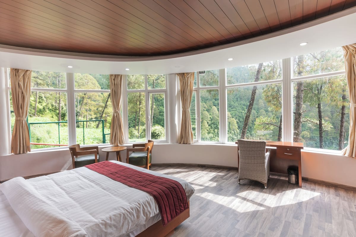 Un dormitorio con una cama grande, un escritorio y una silla, con vistas a árboles a través de amplias ventanas.